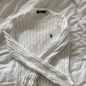Vit stickad tröja från Ralph Lauren - Säljer en stilren vit stickad tröja från Ralph Lauren med kabelstickat mönster och den klassiska loggan på bröstet. Tröjan har långa ärmar och en rund halsringning. Perfekt för en elegant och avslappnad look.