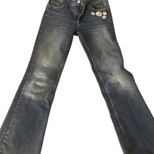 Blå jeans med broderade detaljer - Snygga blå jeans med unika broderade blommor på fram- och baksida. De har en klassisk femficksdesign och en bekväm passform. Perfekta för att ge din outfit en personlig touch.