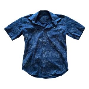 Skjorta från Dolce & Gabbana - Snygg svart skjorta från Dolce & Gabbana med korta ärmar och ett unikt mönster av text och blommor. Skjortan är tillverkad i en bomullsblandning med lite stretch för extra komfort. Perfekt till sommaren.