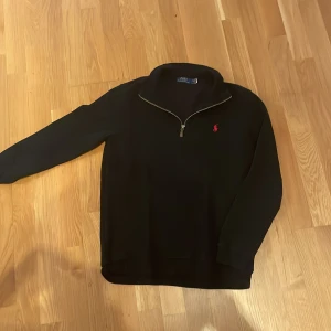 Svart tröja med dragkedja från Polo Ralph Lauren - Säljer en stilren svart tröja från Polo Ralph Lauren med en halv dragkedja och det klassiska röda broderade logotypen på bröstet. Perfekt för en avslappnad men ändå elegant look.