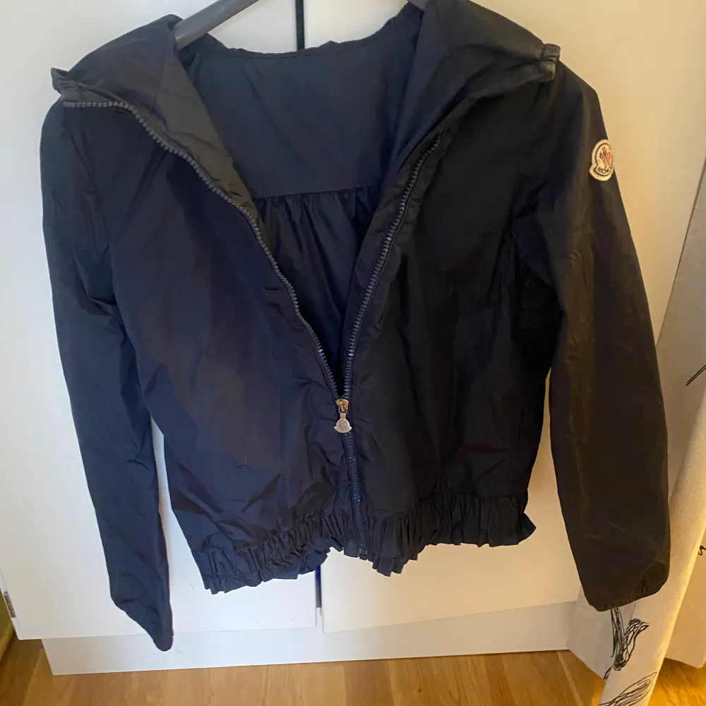 Säljer en snygg svart jacka från Moncler med huva och dragkedja. Jackan har en elastisk nederkant och Moncler-logga på ärmen. Perfekt för kyligare dagar.. Takit.