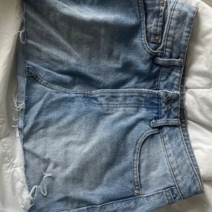 Blå jeansshorts med fransar - Säljer ett par klassiska blå jeansshorts med fransar i nederkant. Perfekta för sommardagar och festivaler. De har en normal passform och är tillverkade i slitstarkt denim. Passar utmärkt till en avslappnad stil.