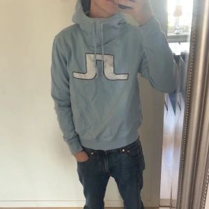 Ljusblå hoodie från J.Lindeberg - Säljer en snygg ljusblå hoodie från J.Lindeberg med deras ikoniska logga på bröstet. Tröjan har en justerbar huva med snörning och långa ärmar. Perfekt för en avslappnad stil.