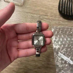 Silverfärgad klocka med fyrkantig urtavla - Elegant klocka med en fyrkantig urtavla och silverfärgad länkarmband. Perfekt som en stilren accessoar för olika tillfällen.