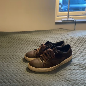 Bruna sneakers från Lanvin - Snygga bruna sneakers från Lanvin med snörning och rund tå. Skorna har en vit sula som ger en fin kontrast till det bruna skinnet. Perfekta för en stilren look.