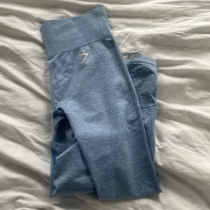 Blå leggings från Gymshark - Snygga blå leggings från Gymshark med hög midja och ribbad detalj vid midjan. De har en tight passform och är perfekta för träning. Materialet är syntetiskt och har en texturerad yta som ger en cool look. Perfekta för att hålla dig bekväm och stilren under träningspasset.