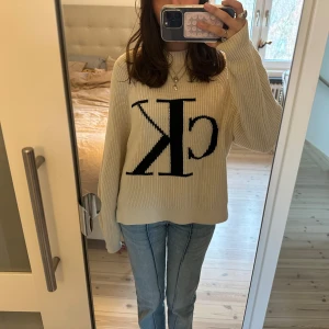 Beige stickad tröja från Calvin Klein - Säljer en snygg beige stickad tröja från Calvin Klein med stor CK-logga i svart på framsidan. Tröjan har långa ärmar och en bekväm passform, perfekt för kyligare dagar. Ett stilrent plagg som passar till det mesta!🌸❤️