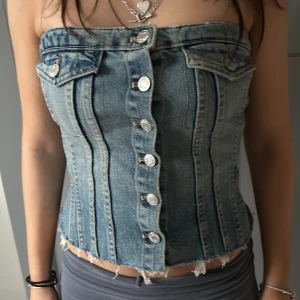 Jeans korsett med knappar - Säljer en cool jeans korsett med knappar framtill och råa kanter. Den har en unik design med fickdetaljer och en tight passform. Perfekt för att ge en edgy look till din outfit.