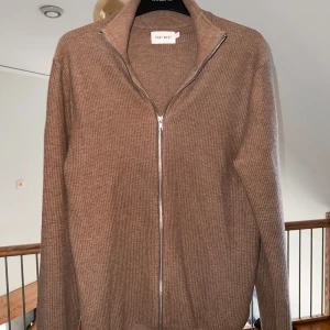Beige hel zip kofta från East West - Säljer en stilren beige kofta från East West med dragkedja framtill. Den är ribbad och har långa ärmar, perfekt för kyligare dagar. Passar både till jeans och mer uppklädda tillfällen.