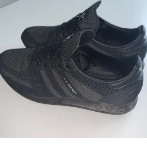 Svarta Adidas L.A. Trainer sneakers Strl 40.5 - Snygga svarta Adidas L.A. Trainer sneakers med klassisk design och tre ränder på sidan. Skorna har en stickad ovandel och snörning för en bekväm passform. Perfekta för både träning och vardag.