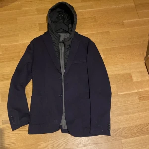 Mörkblå kavajjacka med huva - Snygg mörkblå kavaj med en unik design som inkluderar en avtagbar huva. Kavajen har knappar framtill och är perfekt för en stilren look. Passar bra till både vardag och mer formella tillfällen.