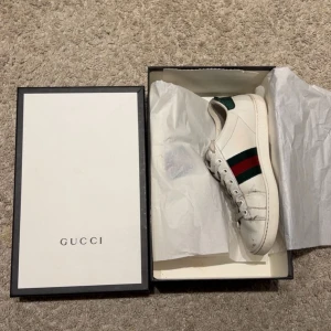Vita sneakers från Gucci med broderad detalj - Snygga vita sneakers från Gucci med klassiska gröna och röda ränder samt en broderad bi-detalj på sidan. Skorna har snörning och en unik design med röd och grön häl i ormskinnsmönster. Perfekta för att ge din outfit en lyxig touch.