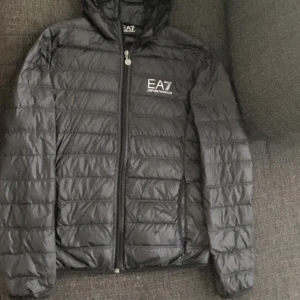 Svart dunjacka från Emporio Armani - Snygg svart dunjacka från Emporio Armani EA7 med dragkedja framtill. Jackan har en quiltad design och en praktisk huva. Perfekt för kyligare dagar och ger en stilren look.