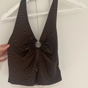 Brun mönstrad topp från h&m - Snygg brun topp från h&m med ett unikt mönster och djup v-ringning. Toppen har en ringdetalj framtill som ger en cool look. Perfekt för en kväll ute eller en avslappnad dag.