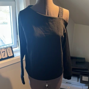 Svart oneshoulder tröja från KappAhl med glitter band på ena sidan  - Snygg svart oneshoulder tröja från KappAhl med glittrig axelrem. Perfekt för en stilren look med en twist. Tröjan har långa ärmar och en bekväm passform. Stl. 158-164