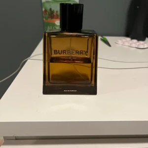 Burberry Eau de Parfum - Elegant och tidlös parfym från Burberry i en stilren, fyrkantig flaska med mörk bärnstensfärg. Flaskan har en svart kork och den klassiska Burberry-loggan på framsidan. Perfekt för den som söker en sofistikerad doftupplevelse.