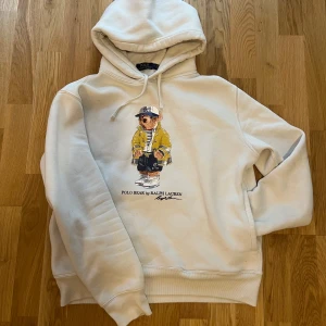Vit hoodie från Ralph Lauren  - Säljer en vit hoodie från Ralph Lauren med ett coolt björnmotiv på framsidan. Hoodien har en klassisk design med justerbar huva och ribbade muddar vid ärmslut och nederkant. Perfekt för en avslappnad stil!
