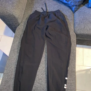 Svarta mjukisbyxor från Adidas - Säljer ett par svarta mjukisbyxor från Adidas med elastisk midja och snörning. Byxorna har en avslappnad passform och ribbade muddar vid bensluten. Adidas-loggan syns längs benet i vitt.