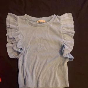 Ljusblå ribbad topp med volangärmar från Zara - Säljer en söt ljusblå ribbad topp från Zara med volangdetaljer på ärmarna. Perfekt för en stilren och trendig look. Toppen har en rund halsringning och korta ärmar.