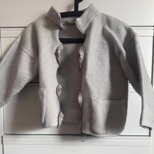 Grå cardigan jacka med volangdetaljer och trekvartsärmar - Säljer en stilren grå cardigan jacka med unika volangdetaljer längs dragkedjan. Jackan har en hög krage och långa ärmar, perfekt för kyligare dagar. Den är tillverkad i ett mjukt material och har praktiska fickor framtill. Dragkjedjan är sådär, står M i men passar mer än s