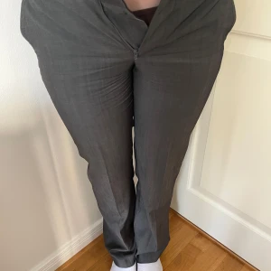 Grå kostymbyxor från Tiger of Sweden - Snygga grå kostymbyxor från Tiger of Sweden med en klassisk passform. Byxorna har en dragkedja framtill och är perfekta för en stilren look. De är tillverkade i ett elegant material som ger en sofistikerad känsla.