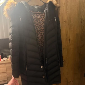 Svart dunjacka med pälsdetaljer från Hollies - Snygg svart dunjacka från Hollies med pälsdetaljer. Jackan har dragkedja och knappar framtill samt en leopardmönstrad insida. Perfekt för kalla dagar! Original pris = ≈ 5000kr