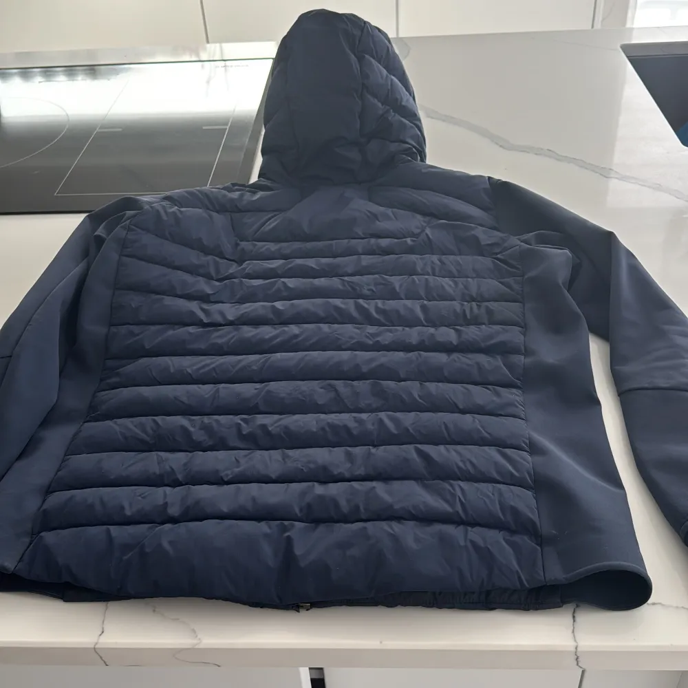 Säljer en blå pufferjacka från Sail Racing med huva och dragkedja. Jackan har ett broderat märke på bröstet och är perfekt för kyligare väder. Den är både stilren och funktionell.. Takit.