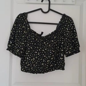 Svart blommig topp från Divided - Säljer en söt svart blommig topp från Divided. Toppen är croppad med korta ärmar och har ett elastiskt band vid halsen och midjan för en bekväm passform. Perfekt för en somrig look! 🌼