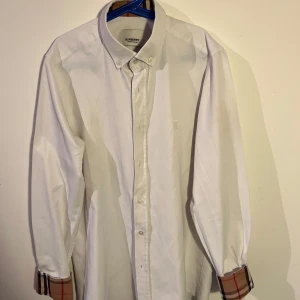 Vit skjorta från Burberry - Snygg vit skjorta i storlek S från Burberry med klassiska rutiga detaljer på ärmsluten. Qr kod finns och skjortan har en broderad logotyp på bröstet och knappar framtill. Perfekt för en stilren look till sommaren. Skjortan är i 9/10 skick och passar någon mellan 160-175 cm och 55-65 kg😇😇😇