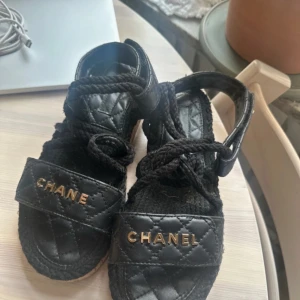 Svarta sandaler från Chanel - Snygga svarta sandaler från Chanel med quiltad design och guldiga detaljer. Sandalerna har en bred rem med logotyp och snörning.  Det saknas ett L men kan lätt fixas genom Chanel garanti! 