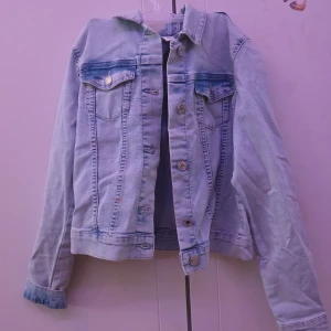 Ljusblå jeansjacka - Snygg ljusblå jeansjacka med klassisk design och knappar framtill. Jackan har två bröstfickor med lock och knappdetaljer. Perfekt för en avslappnad stil.