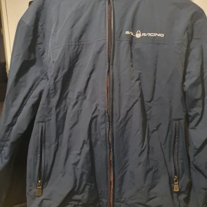 Blå vindjacka från Sail Racing - Säljer en blå vindjacka från Sail Racing med dragkedja framtill och två fickor med dragkedjor. Jackan har en stilren design med logotyp på bröstet och ryggen. Perfekt för blåsiga dagar.
