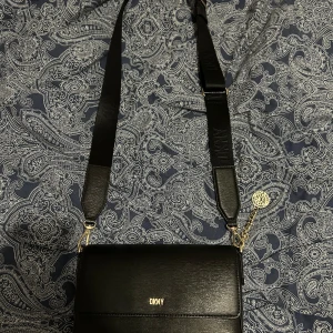 Svart axelväska från DKNY - Snygg svart axelväska från DKNY i skinn med justerbar axelrem. Väskan har en stilren design med guldfärgade detaljer och logotyp framtill. Perfekt för att bära dina nödvändigheter med stil. Pris kan diskuteras 