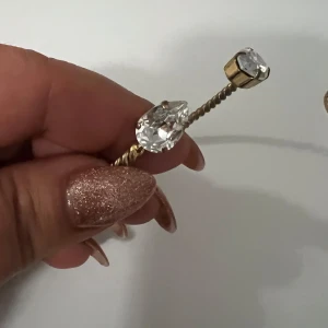 Caroline Svedbom Guldarmband med kristaller - Elegant guldarmband med en tvinnad design och stora klara kristaller i ändarna. Perfekt för att ge en glittrig touch till din outfit.