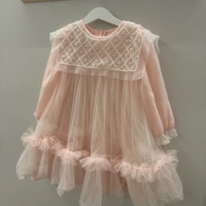 Rosa klänning med pärlor 3-5 år  - Söt rosa klänning med volanger och pärldetaljer på bröstet. Den har långa ärmar och en luftig, flödande design som ger en drömlik känsla. Perfekt för speciella tillfällen.
