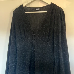 Svart glittrig klänning  - Snygg svart klänning från Ginatricot med glittrigt material och knappar framtill. Den har en v-ringad design och långa ärmar, perfekt för en kväll ute. Klänningen har en elegant och stilren look. Sitter väldigt fint på kroppen, liknar Adoore klänningarna. 