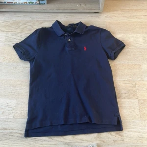 Mörkblå pikétröja från Ralph Lauren - Säljer en stilren mörkblå pikétröja från Ralph Lauren med röd broderad logga på bröstet. Tröjan har korta ärmar och en klassisk krage med knappar. Perfekt för en avslappnad men ändå snygg look. Passar 12 åringar 