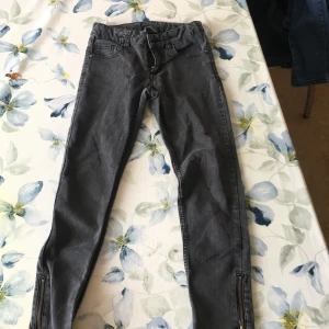 Svart/grå jeans med dragkedja - Snygga svart/grå jeans med tight passform och dragkedjor vid bensluten. De har en klassisk femficksdesign och en dragkedja i gylfen. Jeansen är stretchiga och sköna. Perfekta för en stilren look.