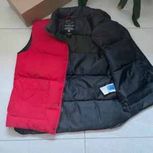 Svart och röd dunväst från Canada Goose - Säljer en snygg svart och röd dunväst från Canada Goose. Västen har en broderad vit lönnlöv på framsidan och är perfekt för kyliga dagar. Den har en dragkedja och knappar för extra skydd mot väder och vind. MVH EliteVolt