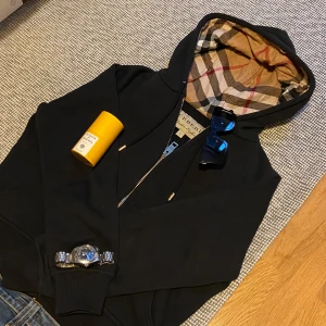 Svart hoodie från Burberry - Säljer en stilren svart hoodie från Burberry med klassiskt rutigt mönster i huvan. Tröjan har en dragkedja framtill och är perfekt för en avslappnad look. Passar bra till både jeans och sneakers.