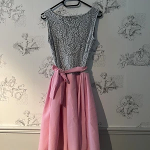 Rosa och grå spetsklänning med rosett - Supersöt klänning med grått spetstopp och vid rosa kjol. Midjan markeras av ett brett band med rosett. Ärmlös modell med rund halsringning, perfekt för speciella tillfällen. Storlek S