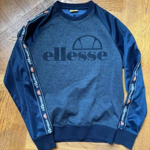 Blå sweatshirt från Ellesse med logga - Snygg blå sweatshirt från Ellesse med stor logga framtill och logoband längs ärmarna. Tröjan har rund hals, långa ärmar och ribbade muddar. Perfekt för en sportig och avslappnad stil.