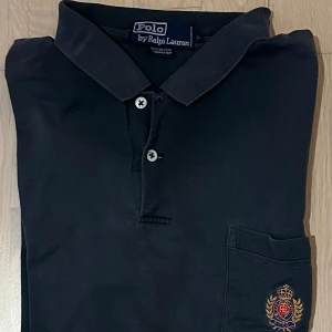Svart pikétröja från Polo Ralph Lauren - Klassisk svart pikétröja från Polo Ralph Lauren i 100% bomull. Tröjan har korta ärmar, krage med knappar och en bröstficka med broderad logga i guld och rött. Perfekt för en stilren och tidlös look.
