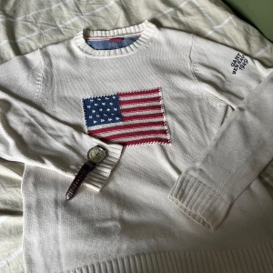 Vit stickad tröja från GANT med flagga - Säljer en vit stickad tröja från GANT med broderad amerikansk flagga på bröstet och texten 'GANT NEW HAVEN 1949' på ärmen. Tröjan har rund halsringning, ribbade muddar och är tillverkad i 100% bomull. Perfekt för dig som gillar klassisk preppy-stil.