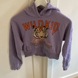 Lila hoodie med tigertryck från Lindex - Cool lila hoodie från Lindex med tryck av en tiger och texten 'WILD KID' i orange. Tröjan är croppad, har huva och långa ärmar. Perfekt för dig som gillar streetstyle och vill sticka ut med ett djurmotiv.