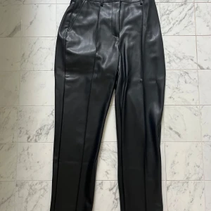 Svarta kostymbyxor i skinnimitation NA-KD - Snygga svarta kostymbyxor från NA-KD i skinnimitation. Byxorna har raka ben, pressveck och hällor för bälte. De har fickor fram och bak samt stängs med dragkedja och hyska. Perfekta för en edgy och trendig look.