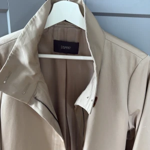 Beige trenchcoat från Esprit, M - Snygg beige trenchcoat från Esprit med klassisk krage, knappar vid ärmslut och dragkedja framtill. Jackan har två stora fickor och ett bälte i midjan för extra stilpoäng. Perfekt för dig som gillar en clean och tidlös look.