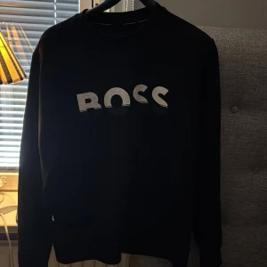 Svart sweatshirt från BOSS med tryck - Svart sweatshirt från BOSS i storlek S med stort BOSS-tryck i vitt på bröstet. Tröjan har rund hals och långa ärmar, perfekt för en clean och stilren look. Materialet känns mjukt och skönt mot huden, och passformen är normal.