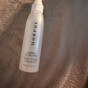 Morphe Luminous Setting Spray - Säljer en Morphe Luminous Setting Spray i en snygg, pärlemorskimrande plastflaska med svart text. Sprayen ger en glowig finish och håller sminket på plats hela dagen. Flaskan rymmer 125 g och har en praktisk sprayfunktion för enkel applicering.