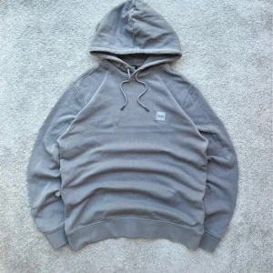 Mörkgrå Hugo Boss hoodie - Säljer en stilren grå hoodie från BOSS med broderad logotyp på bröstet. Tröjan har huva med snörning och långa ärmar. Mjuk bomullskvalitet och relaxed passform gör den perfekt för chill dagar. Enkelt och cleant plagg som funkar till jeans eller joggers. Den är helt ny bara att jag inte har original förpackningen. Skriv vid frågor:)